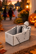 Small skeleton in a coffin-shaped treat holder on a Halloween porch with trick-or-treaters in the background